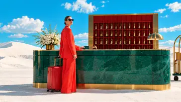 Una donna in un tailleur rosso con una valigia rossa, in piedi alla reception di marmo verde di un albergo, tra dune di sabbia bianca